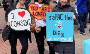 Three students hold signs for SAPAC the Diag event