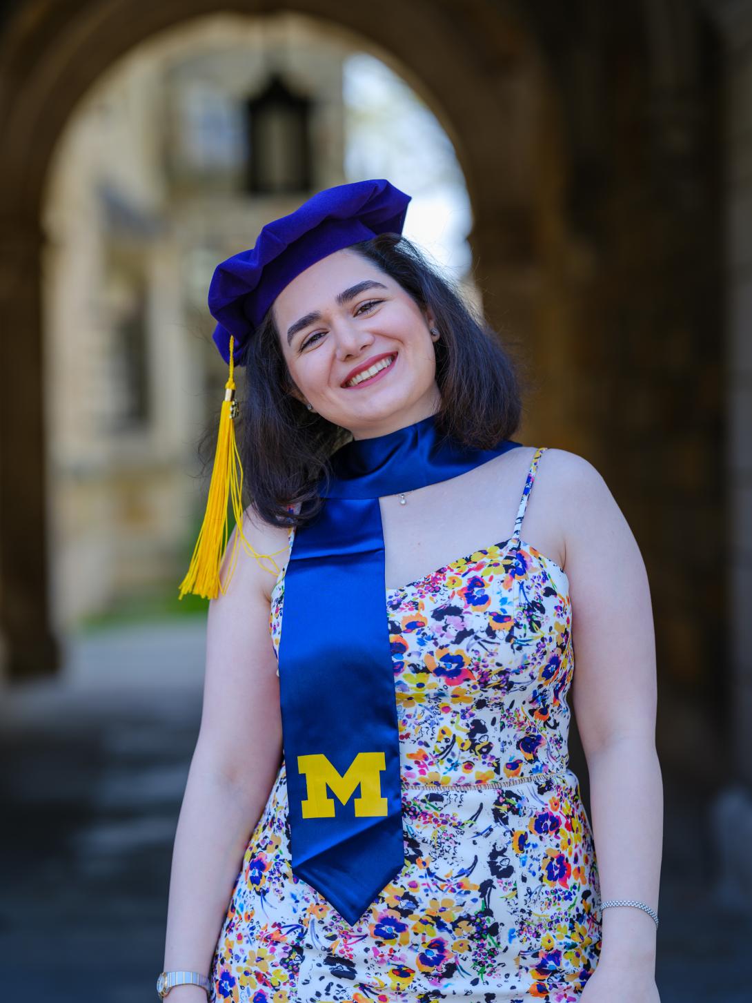 Reyhaneh Najafikoupaei is a woman with dark hair posting with a Michigan Graduation cap and stole in a floral dress.